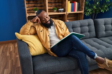 Young african american man reading book sitting on sofa at home