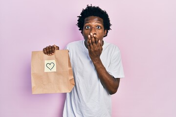Young african american man holding take away paper bag with heart reminder covering mouth with...