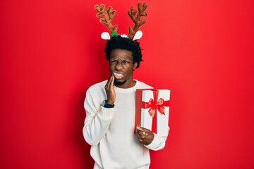 Young african american man wearing deer christmas hat holding gift touching mouth with hand with...