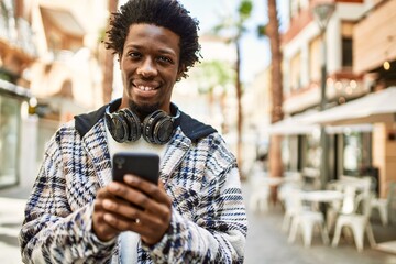 Handsome black man with afro hair wearing headphones smiling happy outdoors using smartphone