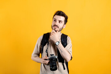 Handsome caucasian man wearing beige t-shirt posing isolated over yellow background serious face thinking about question, very confused idea while holding a dslr camera.