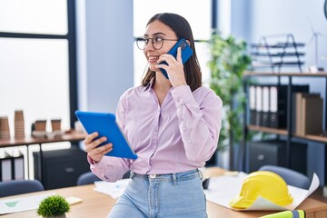 Young hispanic woman architect talking on the smartphone holding touchpad at office