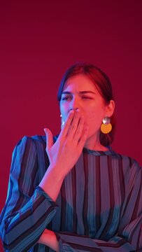 Bored woman. Time waste. Sleep deprivation. Tired yawning girl with apathy expression in neon light on ruby red background vertical.
