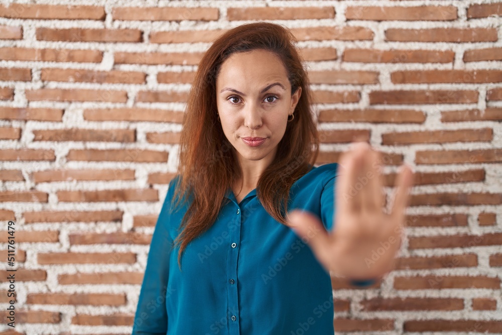 Poster brunette woman standing over bricks wall doing stop sing with palm of the hand. warning expression w