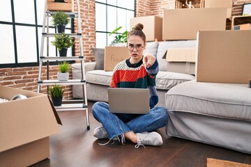 Young hispanic girl sitting on the floor at new home with laptop pointing with finger to the camera and to you, confident gesture looking serious