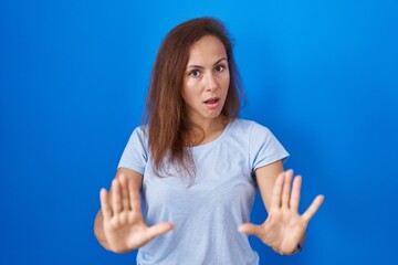 Brunette woman standing over blue background moving away hands palms showing refusal and denial...