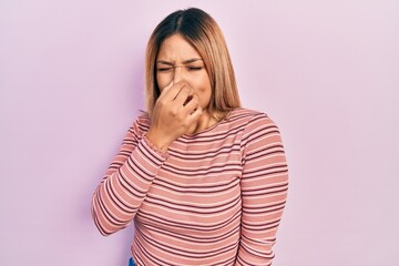 Beautiful hispanic woman wearing casual striped sweater smelling something stinky and disgusting, intolerable smell, holding breath with fingers on nose. bad smell - obrazy, fototapety, plakaty