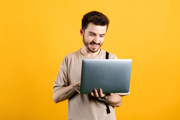 Caucasian man wearing beige tee posing isolated over yellow background standing and using laptop pc computer smiling with a happy and cool smile on face. Showing teeth.