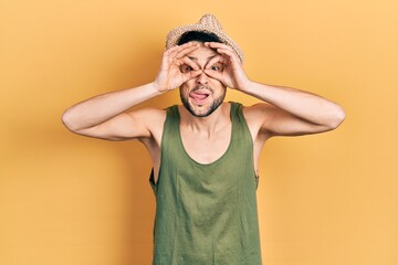 Young hispanic man wearing summer hat doing ok gesture like binoculars sticking tongue out, eyes...