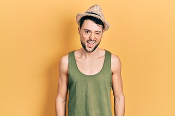 Young hispanic man wearing summer hat winking looking at the camera with sexy expression, cheerful...