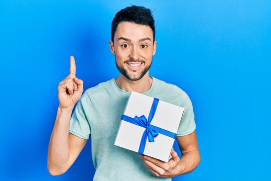 Young hispanic man holding gift smiling with an idea or question pointing finger with happy face, number one