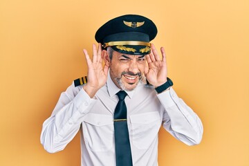 Handsome middle age man with grey hair wearing airplane pilot uniform trying to hear both hands on...