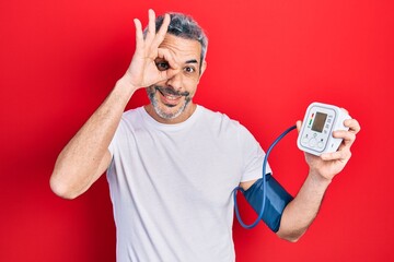 Handsome middle age man with grey hair using blood pressure monitor smiling happy doing ok sign...