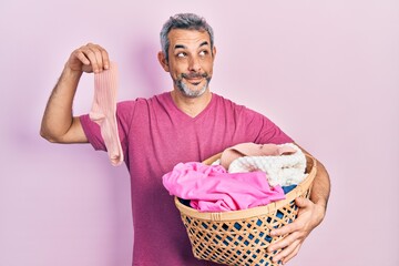 Handsome middle age man with grey hair holding laundry basket and dirty sock smiling looking to the...