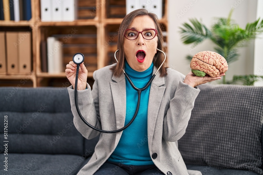 Sticker Middle age hispanic woman working at therapy office holding brain afraid and shocked with surprise and amazed expression, fear and excited face.