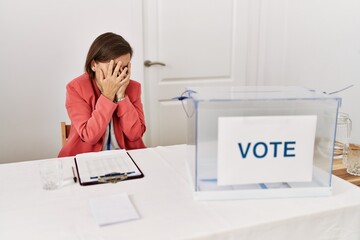 Beautiful middle age hispanic woman at political election sitting by ballot with sad expression covering face with hands while crying. depression concept.