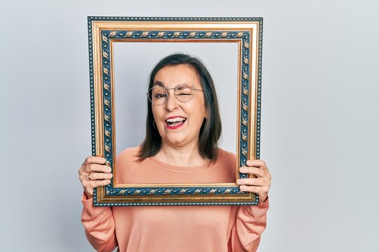 Middle age hispanic woman holding empty frame winking looking at the camera with sexy expression, cheerful and happy face.