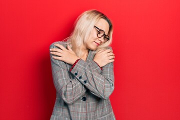 Fototapeta premium Beautiful caucasian blonde woman wearing business jacket and glasses hugging oneself happy and positive, smiling confident. self love and self care