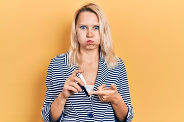 Beautiful caucasian blonde woman holding glucometer device puffing cheeks with funny face. mouth inflated with air, catching air. - obrazy, fototapety, plakaty