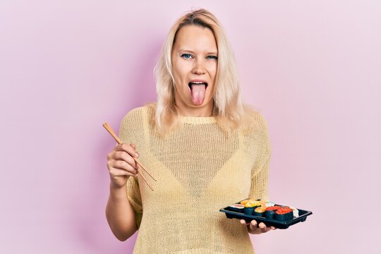 Beautiful caucasian blonde woman eating sushi using chopsticks sticking tongue out happy with funny expression.