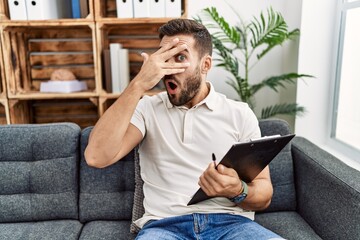 Handsome hispanic man holding clipboard working at psychology clinic peeking in shock covering face and eyes with hand, looking through fingers with embarrassed expression.