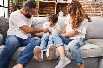 Family doing tickle to son sitting on sofa at home - obrazy, fototapety, plakaty
