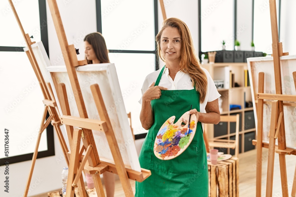 Poster young hispanic middle age woman at art classroom pointing finger to one self smiling happy and proud