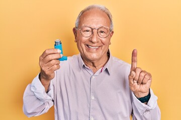 Senior man with grey hair holding medical asthma inhaler surprised with an idea or question pointing finger with happy face, number one - obrazy, fototapety, plakaty