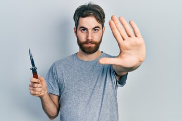 Caucasian man with beard holding pocket knife with open hand doing stop sign with serious and...