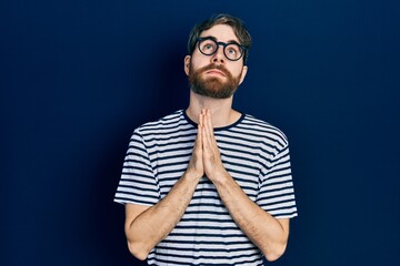 Fototapeta premium Caucasian man with beard wearing striped t shirt and glasses begging and praying with hands together with hope expression on face very emotional and worried. begging.