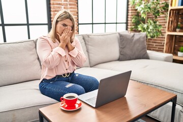 Young hispanic woman using laptop sitting on the sofa at home smelling something stinky and...