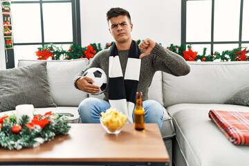 Young hispanic man football hooligan holding ball and beer with angry face, negative sign showing...