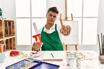 Young hispanic man at art studio holding degree pointing with finger up and angry expression, showing no gesture