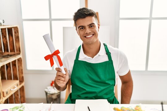 Young hispanic man smiling confident holding diploma at art studio