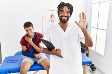 Young hispanic man working at pain recovery clinic with a man with broken arm waiving saying hello happy and smiling, friendly welcome gesture