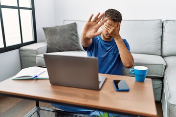 Young handsome hispanic man using laptop sitting on the floor covering eyes with hands and doing stop gesture with sad and fear expression. embarrassed and negative concept.