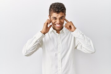 Young handsome hispanic man standing over isolated background covering ears with fingers with annoyed expression for the noise of loud music. deaf concept.