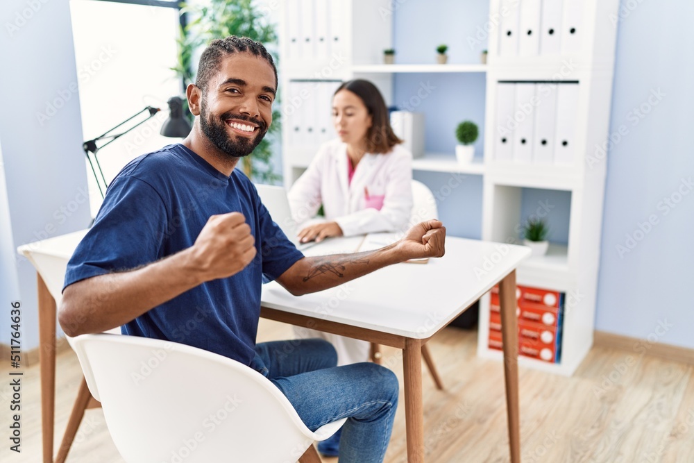 Wall mural Young hispanic man at doctor clinic screaming proud, celebrating victory and success very excited with raised arm