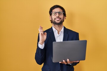 Handsome latin man working using computer laptop gesturing finger crossed smiling with hope and eyes closed. luck and superstitious concept.