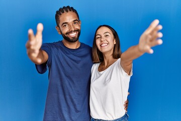 Young hispanic couple standing together looking at the camera smiling with open arms for hug. cheerful expression embracing happiness. - obrazy, fototapety, plakaty