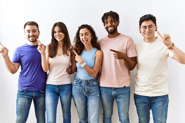 Obraz premium Group of young people standing together over isolated background with a big smile on face, pointing with hand finger to the side looking at the camera.