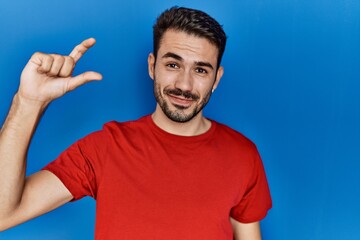 Young hispanic man with beard wearing red t shirt over blue background smiling and confident gesturing with hand doing small size sign with fingers looking and the camera. measure concept.