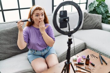 Young redhead woman recording make up tutorial with smartphone at home covering mouth with hand,...