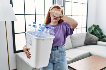 Young redhead woman holding recycling wastebasket with plastic bottles covering eyes with hand, looking serious and sad. sightless, hiding and rejection concept