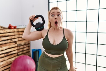 Young redhead woman wearing sportswear using dumbbells scared and amazed with open mouth for...