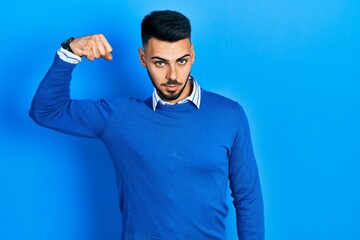 Naklejka premium Young hispanic man with beard wearing casual blue sweater strong person showing arm muscle, confident and proud of power