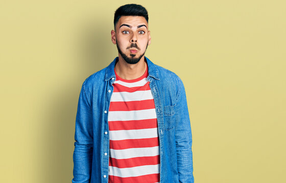 Young hispanic man with beard wearing casual denim jacket puffing cheeks with funny face. mouth inflated with air, crazy expression.