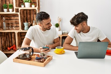 Two hispanic men couple having breakfast using smartphone and laptop at home