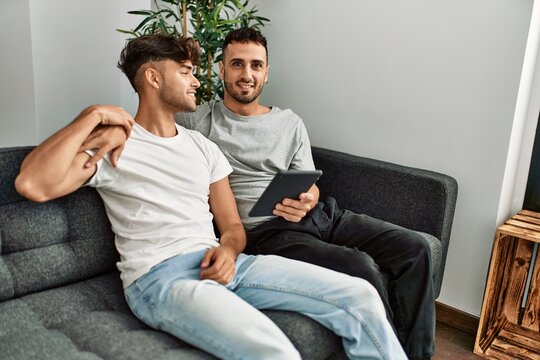 Two hispanic men couple using touchpad hugging each other at home