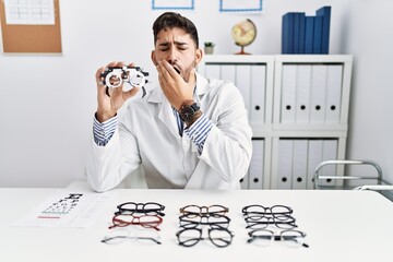 Young optician man holding optometry glasses bored yawning tired covering mouth with hand. restless...